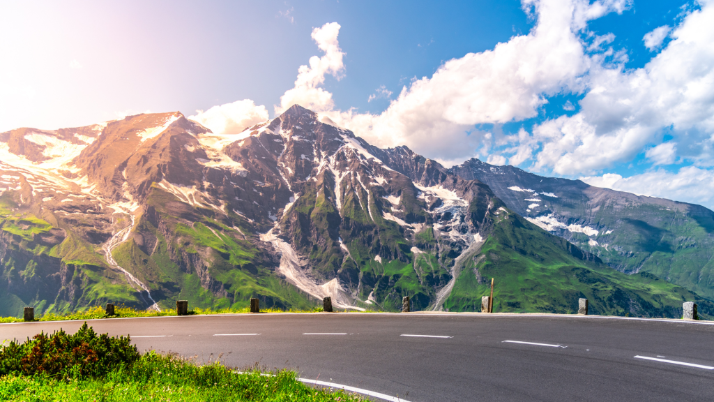 A winding mountain road curves in front of a massive snow-streaked peak under a bright blue sky with scattered clouds