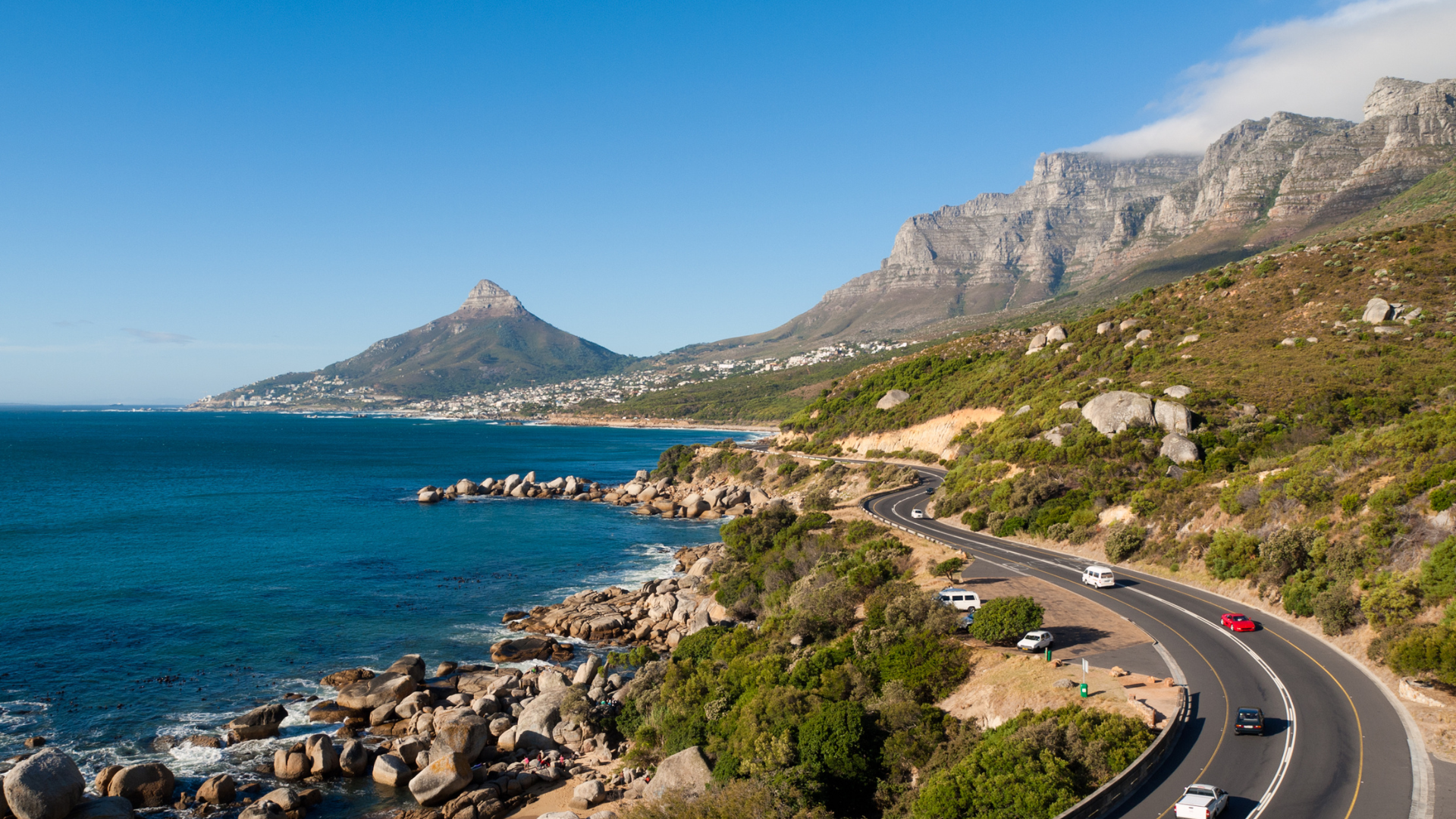 A coastal road curves along rocky cliffs beside deep blue ocean water, with mountains and a small city in the background