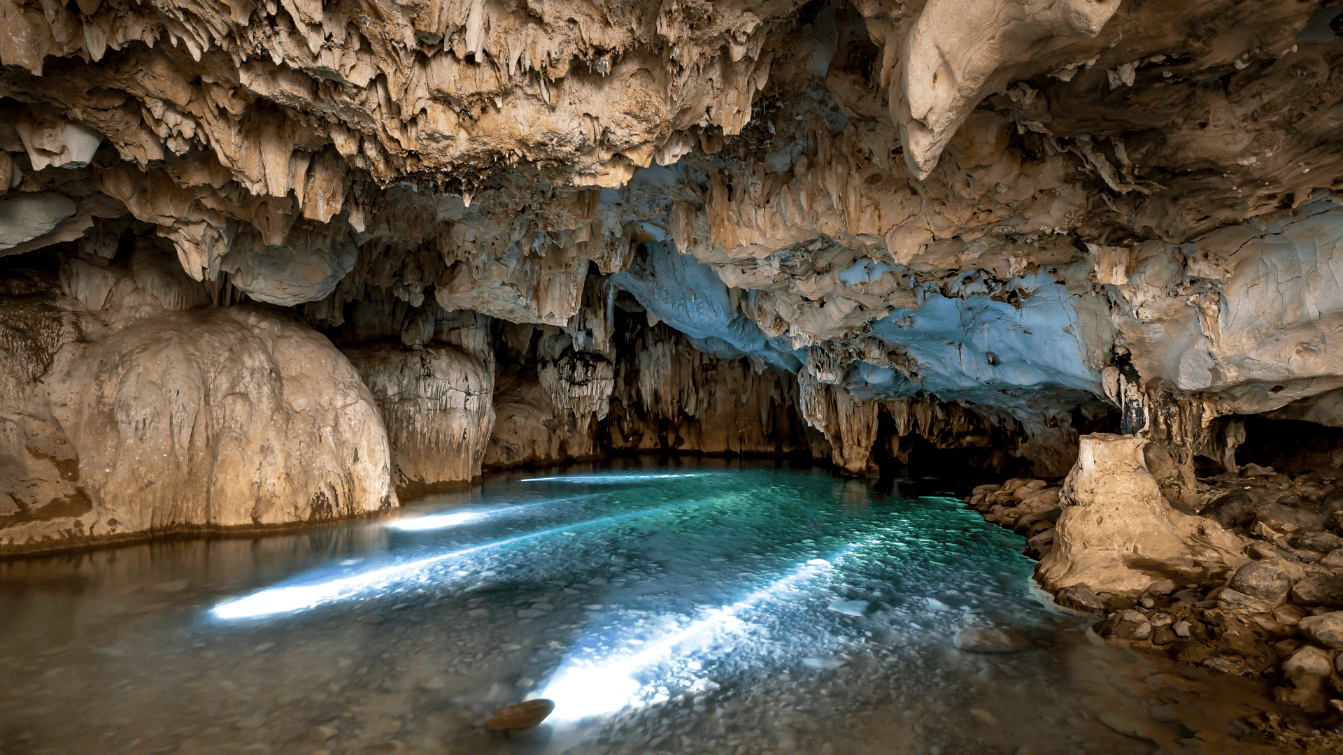 ถ้ำหินงอกหินย้อยสีเบจมีลำธารน้ำใสสะท้อนแสง ทำให้พื้นน้ำเรืองแสงสีฟ้าอ่อน