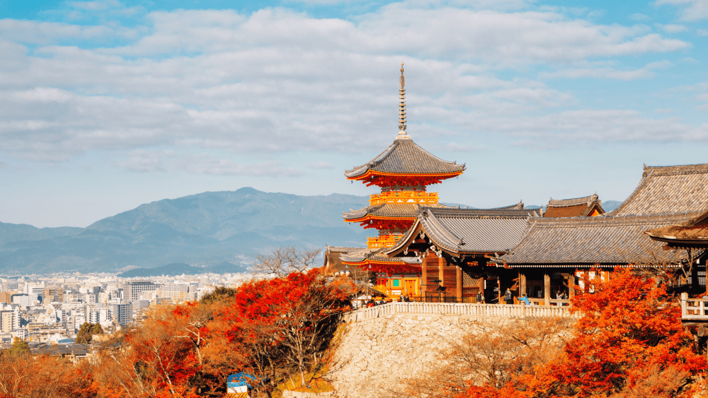 A traditional Japanese temple in Kyoto framed by vivid autumn foliage, with layered pagodas overlooking the city and distant mountains under a soft blue sky.