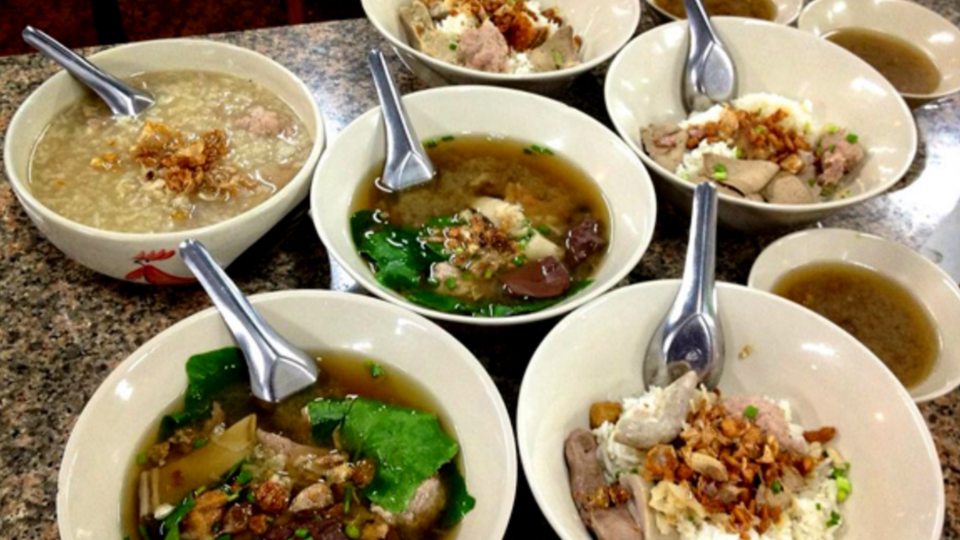An overhead view of several bowls of Asian noodle and soup dishes on a table, topped with sliced meat, greens, fried garlic, and served with metal spoons.