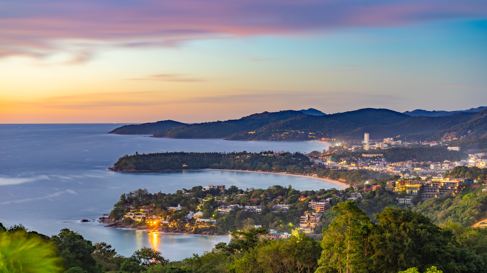 A panoramic view of a coastal city at dusk, with a curved bay, calm sea, green hills, and warm city lights glowing under a pastel sky
