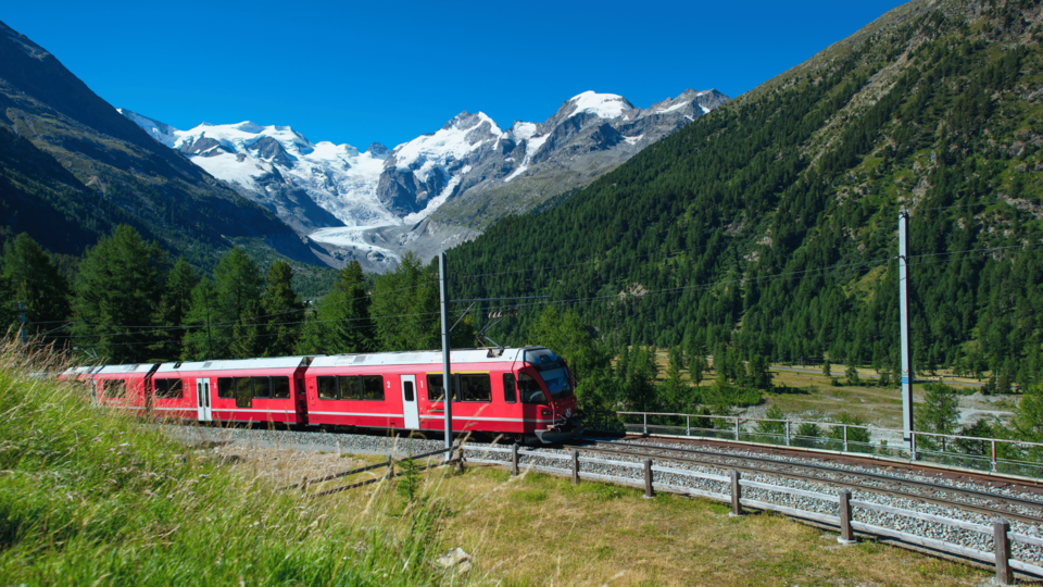 Red scenic train traveling through a green alpine valley with snow-capped mountains