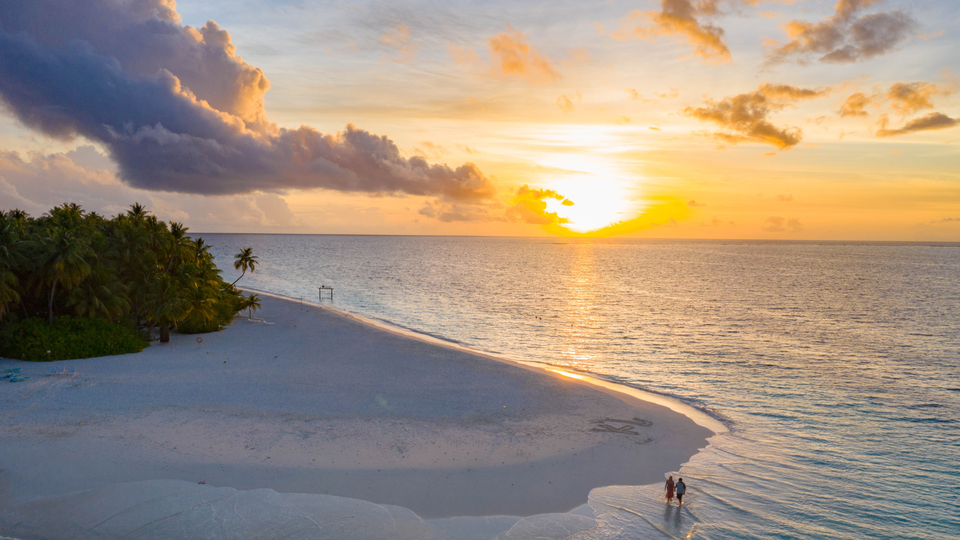 A golden sunset over a quiet tropical beach, with palm trees on the left and two people walking along the shoreline as waves gently roll in.