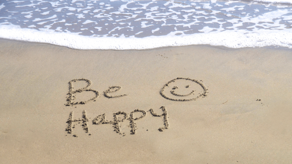The words “Be Happy” written in the sand with a smiley face as waves gently approach the shore