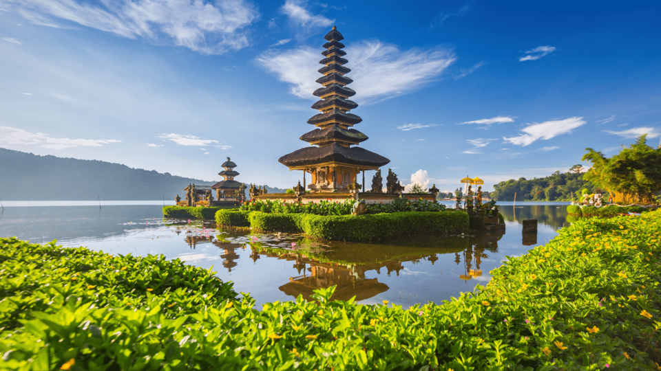 A serene Balinese water temple with tiered pagodas reflected in still lake water, surrounded by vibrant greenery and tropical landscape.