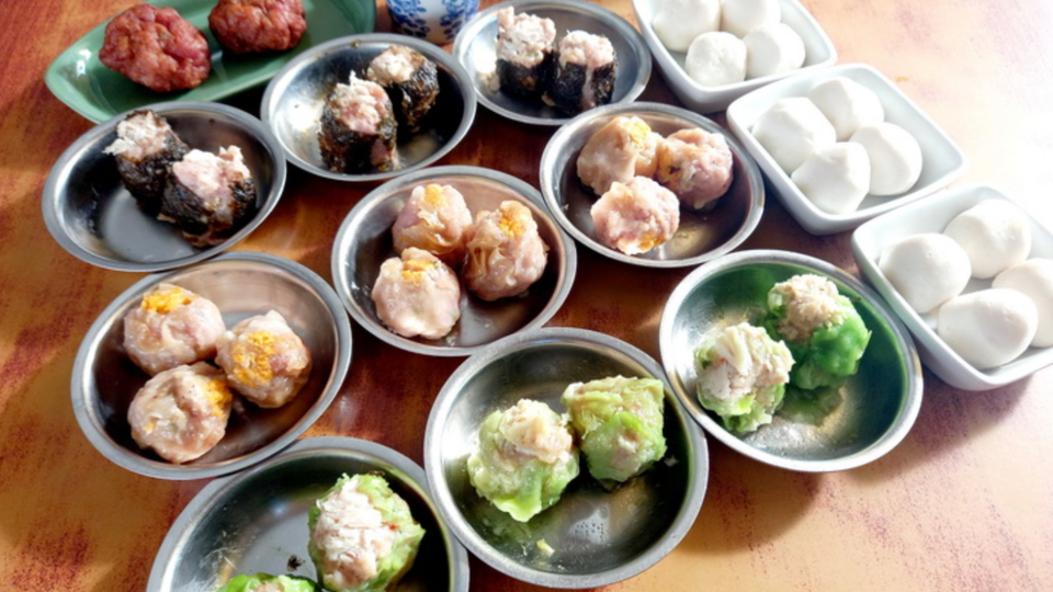 An overhead view of assorted dim sum dishes in small metal and ceramic plates, including shumai, meatballs, rolls, and steamed buns on a wooden table.