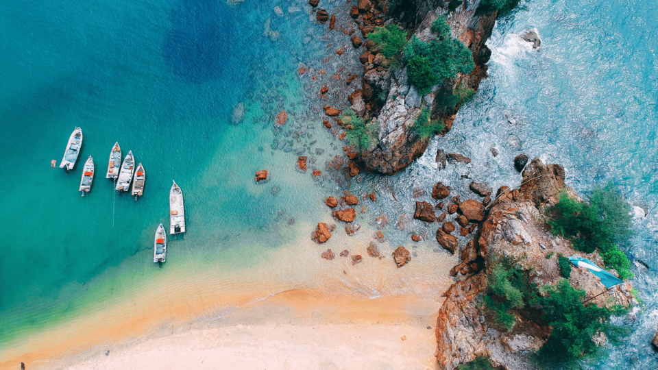 Aerial view of small boats anchored in clear turquoise water near a rocky coastline and sandy beach
