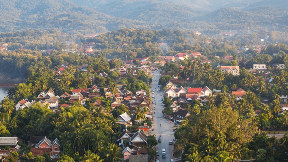 An aerial view of Luang Prabang showing a palm-lined town with traditional rooftops, a central road and forested mountains in the background.