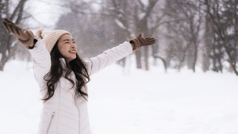 Woman enjoying snowfall in a winter forest, arms raised and smiling
