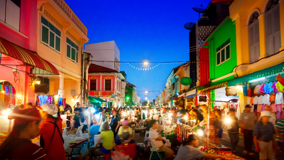 A lively night market street in Phuket, filled with locals and visitors browsing food stalls and shops under warm lights, with colourful historic buildings lining the road at dusk.