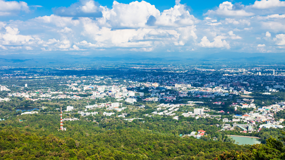 A panoramic view of Chiang Mai city surrounded by lush green hills, dense urban greenery and dramatic cloud-filled blue skies.