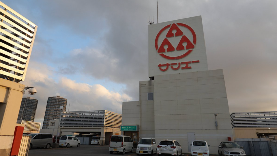 Exterior of a San-A supermarket in Okinawa at dusk, featuring a large red logo on a white building, parked cars in front, and soft evening light in the sky.