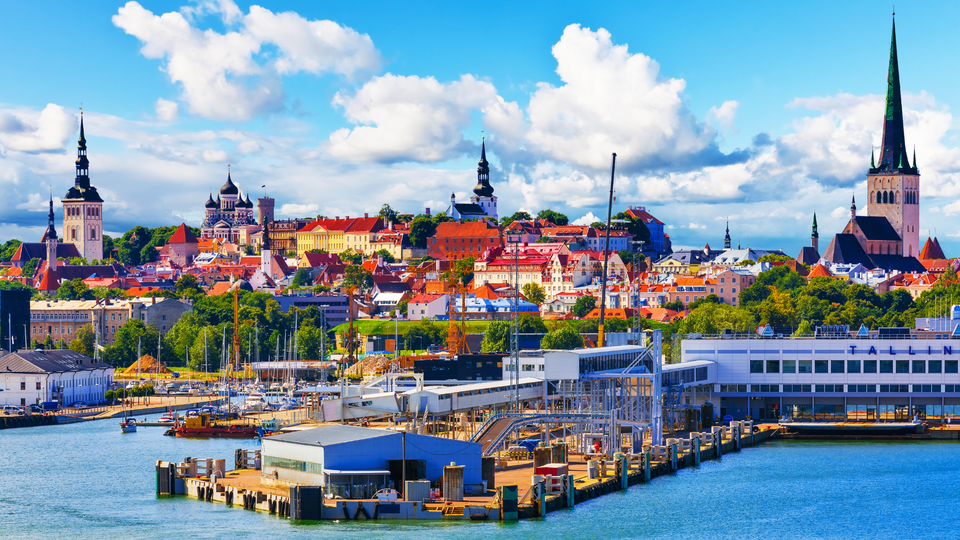 Panoramic view of Tallinn Old Town, Estonia, featuring red rooftops, medieval church spires, and the Baltic Sea harbor under a bright blue sky.