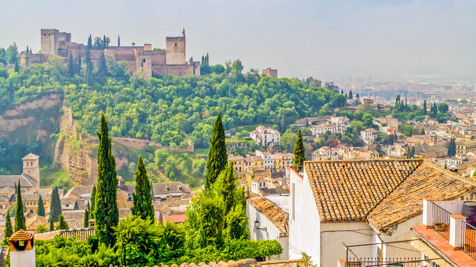 Scenic view of Girona Old Town, Spain, with historic fortress walls on a hill, terracotta rooftops, and lush green cypress trees in a peaceful setting.