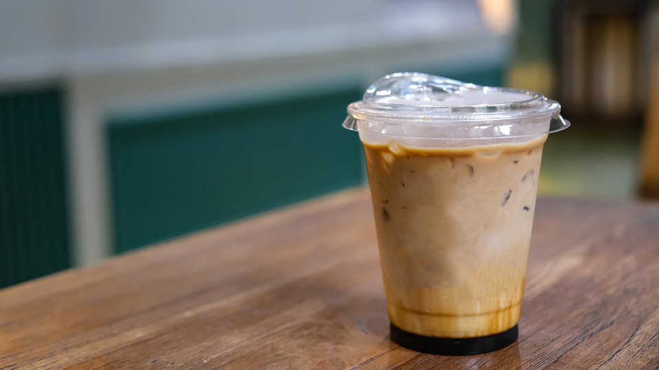 A plastic cup of iced coffee with milk on a wooden table inside a café, with a softly blurred interior in the background.
