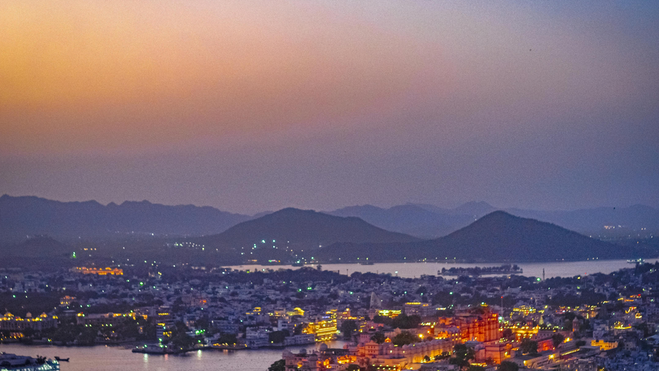Twilight view over a city with glowing lights along the waterfront, mountains layered in the distance, and a soft purple and orange sky at sunset.