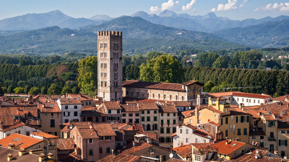 Aerial view of Lucca, Tuscany, Italy, showcasing historic towers, a Romanesque church, and warm terracotta rooftops with mountains in the background.
