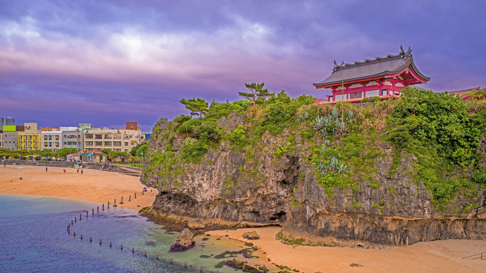 Traditional Okinawan shrine building perched on a rocky green cliff above a sandy beach, with dramatic clouds in the sky and calm ocean water below.