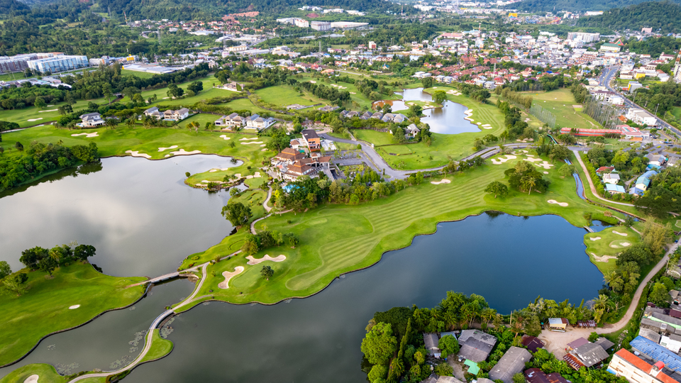 An aerial view of a green golf course in Phuket, with lakes, winding fairways, villas, and residential neighbourhoods surrounded by hills.