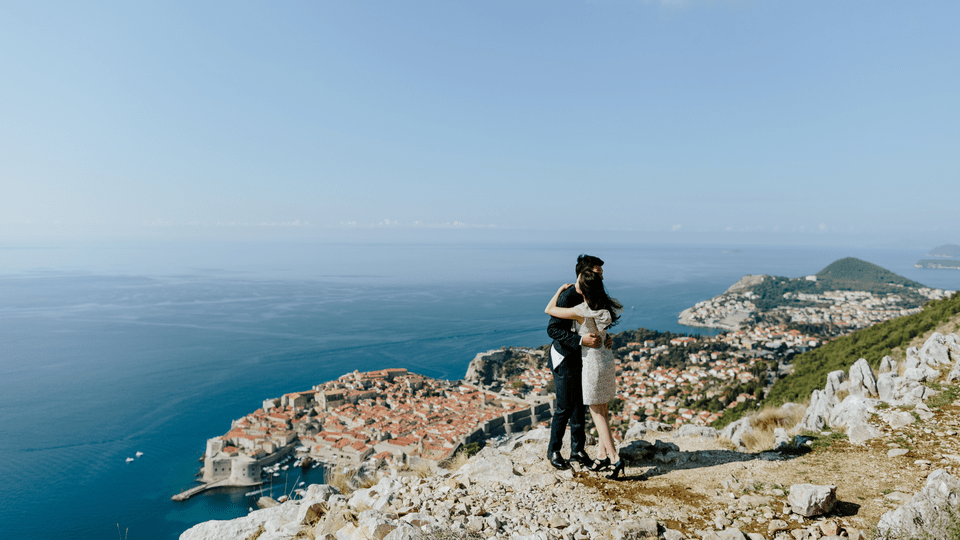 A couple embraces on a rocky hilltop overlooking a coastal city with orange rooftops and clear blue sea stretching to the horizon on a sunny day.