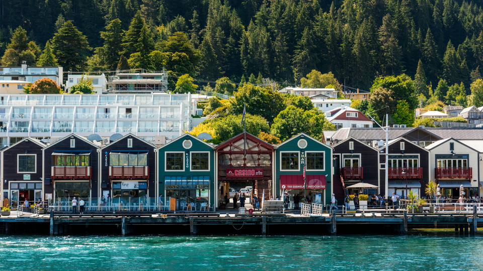 Colorful waterfront buildings and restaurants along Lake Wakatipu with forested mountains in Queenstown, New Zealand.