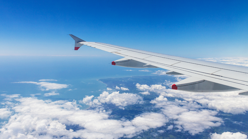 Airplane wing above clouds during flight with blue sky and coastline visible below.